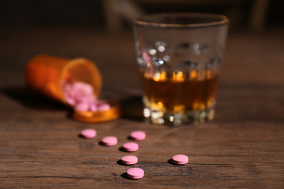 Bottle with pink pills spilling out of it next to a glass with amber colored alcohol.