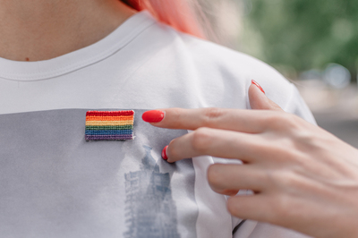 Person pointing to a rainbow flag pin on their shirt.