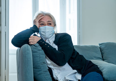 Woman sitting on her couch wearing a protective face mask.